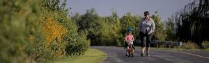 Woman walking beside a young child riding a bike on a paved trail surrounded by trees and yellow wildflowers in Outdoor Sylvania Parks.